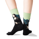 A pair of crew socks worn on a person's legs, featuring a black lower half with a printed Mona Lisa design and a light green upper half with blue mountain graphics.