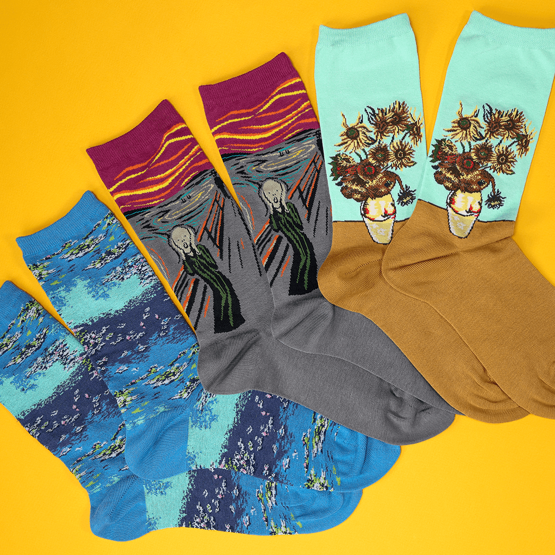 Four pairs of novelty socks featuring famous art prints, arranged in a fan shape on a bright yellow background. The designs include Edvard Munch's The Scream in gray and magenta, Van Gogh's Starry Night in blue, Van Gogh's Sunflowers in light blue, and Van Gogh's Sunflowers in brown.