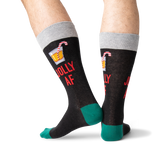 A pair of novelty crew socks worn on a person's legs, featuring a dark charcoal body with a gray cuff, green heels and toes, red text reading 'JOLLY AF', and a small cocktail graphic on the upper calf.
