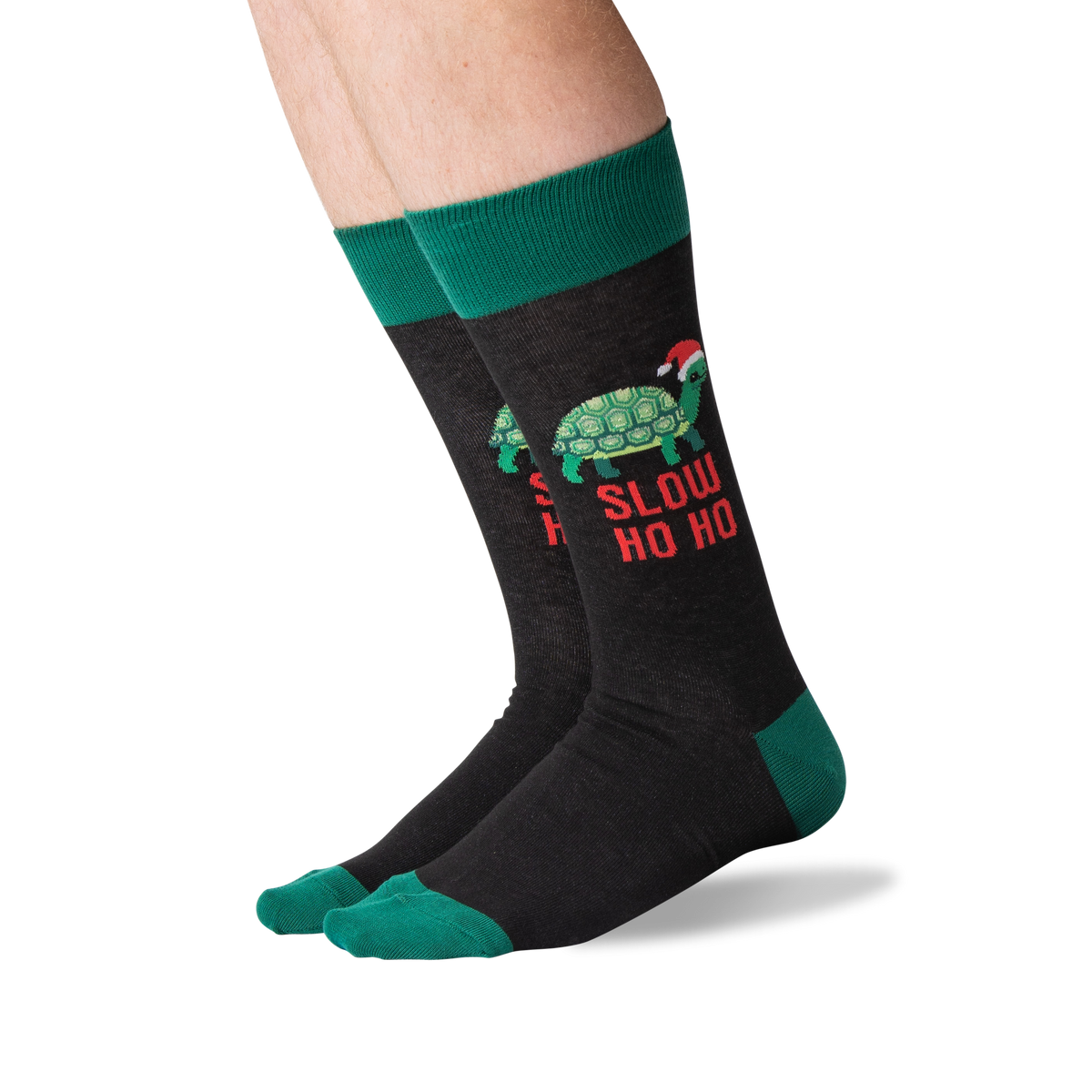 A pair of crew socks worn on a person's legs, featuring a dark charcoal gray body with a green turtle wearing a Santa hat and the text 'SLOW HO HO' in red, plus green cuffs, heels, and toes.