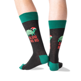 A pair of crew socks worn on a person's legs, featuring a dark charcoal gray body with green cuffs, heels, and toes, and a graphic of a green turtle wearing a red Santa hat with the text 'SLOW HO HO' in red.