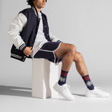 A model sits on a white block wearing a navy and white varsity jacket, matching shorts, white sneakers, and navy crew socks with red stripes and white snowflake patterns.