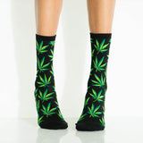 A pair of crew socks with a green cannabis leaf pattern on a black background, worn on a person's legs against a white studio backdrop. thumbnail