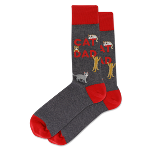A pair of crew socks in dark heather gray with bright red cuffs, heels, and toes. The main body features the words 'CAT DAD' in red, alongside several cartoon cats in gray, white, and yellow.