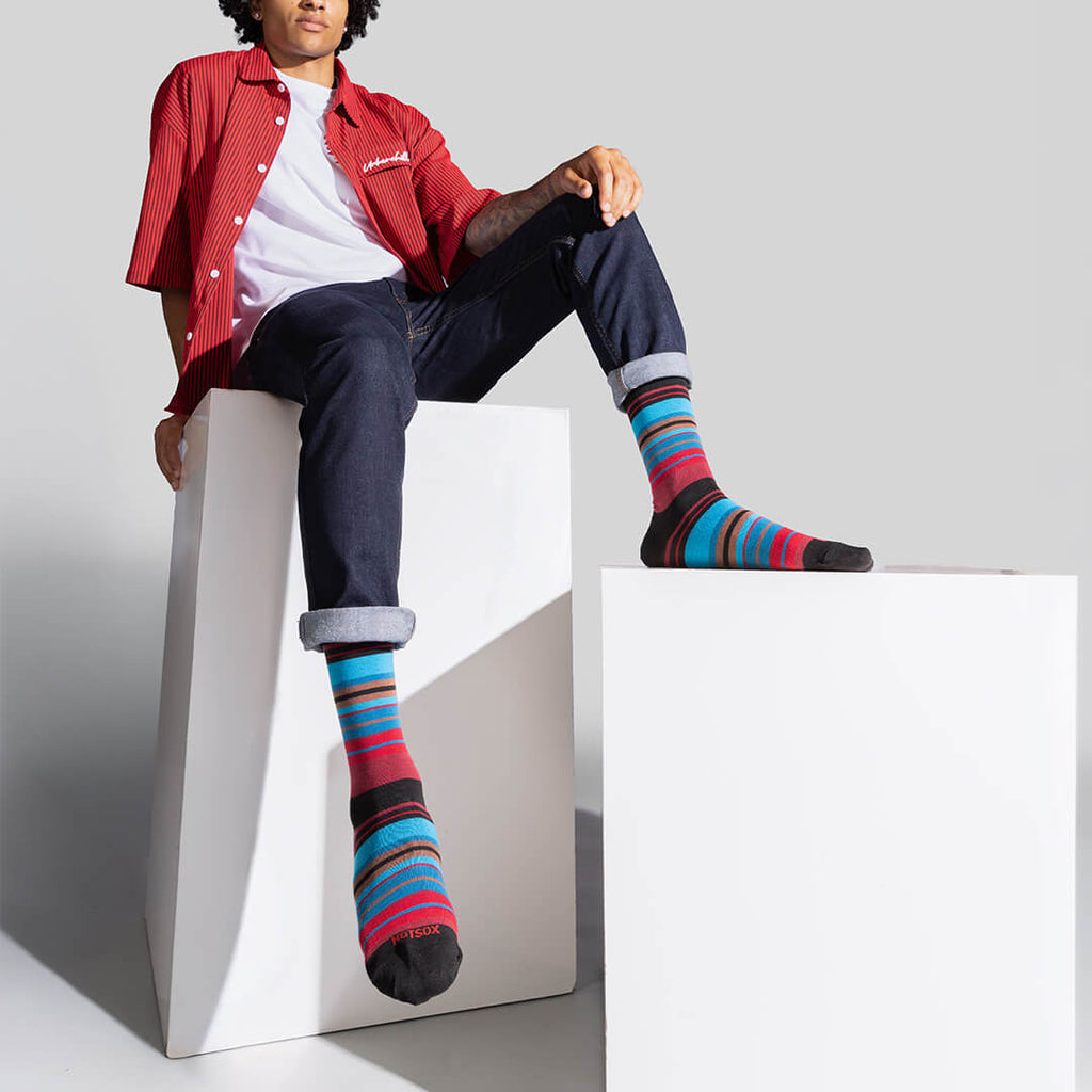A model wearing colorful striped crew socks, a red button-up shirt, and dark jeans sits on white geometric display blocks against a light gray studio background.