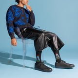 A model sits on a clear acrylic chair wearing a black and blue patterned sweater, black leather-like cropped pants, black sneakers, and black crew socks with colorful graphic prints. The background is a solid light blue studio backdrop.
