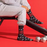 A person seated on a black exercise bench wearing black crew socks with white barbell graphics and red text reading 'PRACTICE SAFE SETS'. The scene is on a bright red studio background with dumbbells nearby. thumbnail