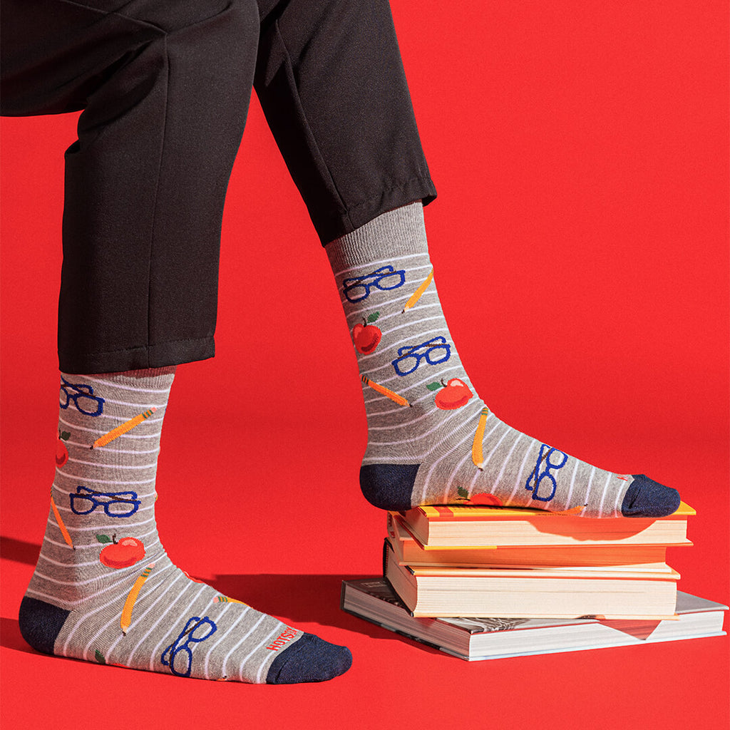 Gray striped socks with a school-themed pattern of blue glasses, yellow pencils, and red apples, worn with black pants and styled on a stack of books against a bright red background.