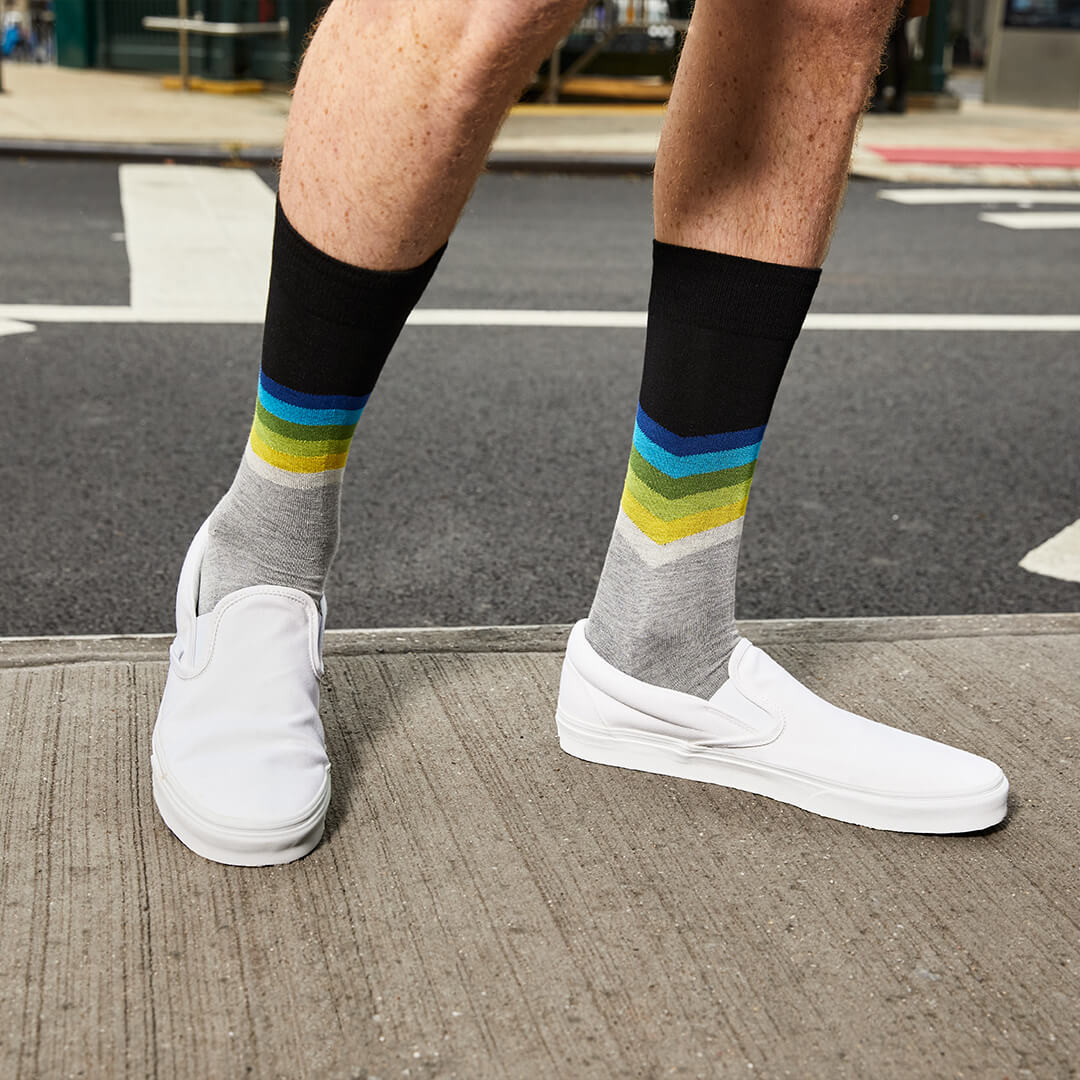 A person wearing gray crew socks with a black top band and multicolor chevron stripes in blue, green, and yellow, paired with white slip-on shoes on a city sidewalk.