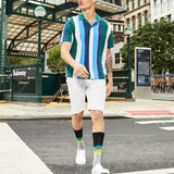 A man walking outdoors in front of a subway station wearing a short-sleeve polo shirt with vertical stripes in green, blue, white, and dark navy, white shorts, black socks with rainbow stripes, and white sneakers. thumbnail