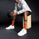Person sitting on a wooden block holding a basketball, wearing gray sweatshirt, black shorts, white sneakers, and orange socks with black stripes. thumbnail