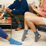Gray ankle socks with a cat face design and pink toes, worn by a person sitting on a couch while playing cards in a cozy living room setting.