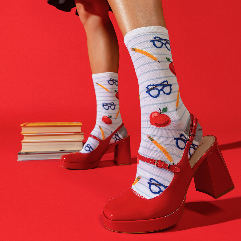 A person wearing white striped socks with red apples, blue glasses, and yellow pencils, paired with red Mary Jane high heels, posed against a solid red background with a stack of books.