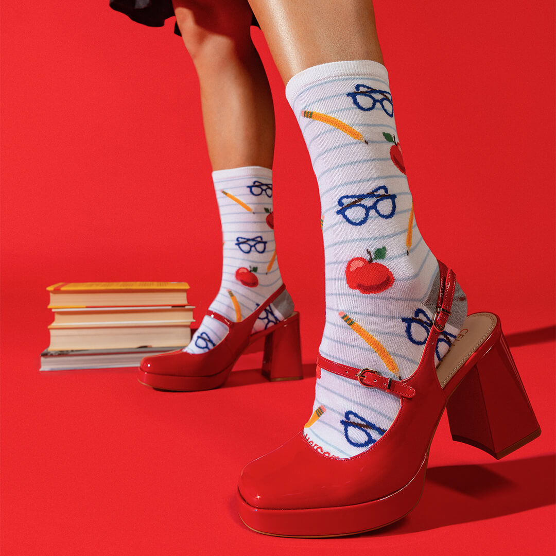 A person wearing white striped socks with red apples, blue glasses, and yellow pencils, paired with red Mary Jane high heels, posed against a solid red background with a stack of books.