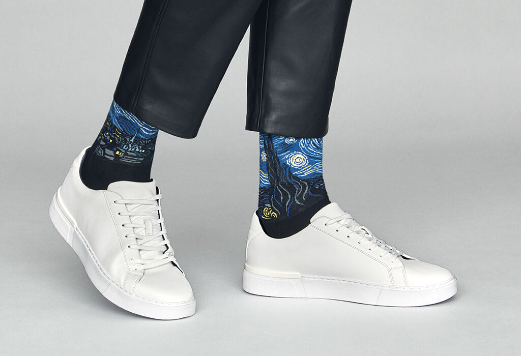 Person wearing black socks with artistic designs and white sneakers on a gray background