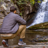 Person in a gray puffer jacket and beige pants sitting by a waterfall.