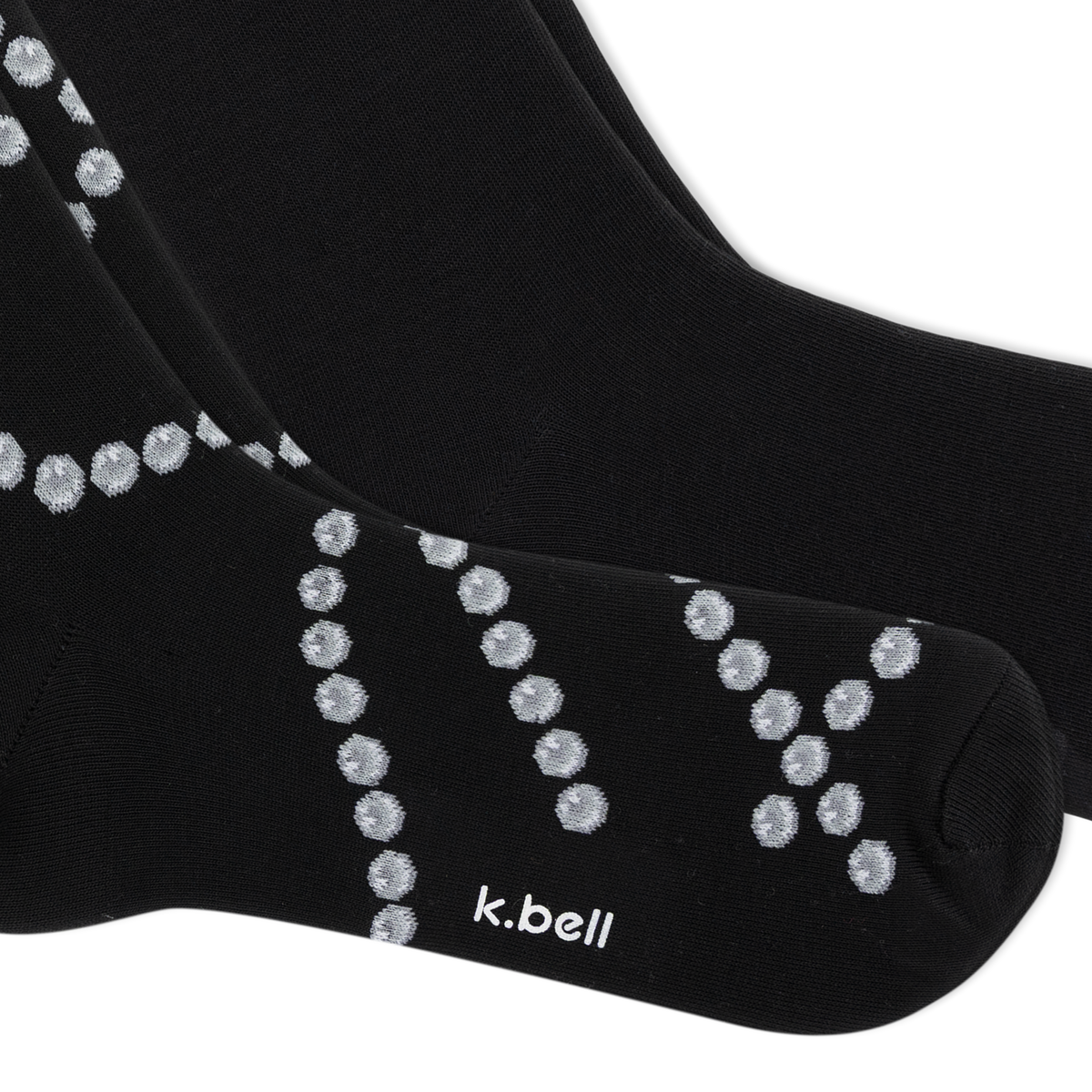 A close-up of a pair of black crew socks with a pattern of small pearl chains, featuring the white text 'k.bell' on the toe area, set against a plain white background.