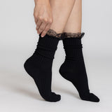 A person wearing black ribbed ankle socks with a black lace ruffle at the top, shown against a light gray studio background.