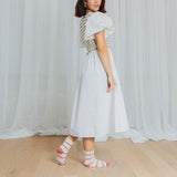 Woman wearing a white dress with a patterned pink and white striped socks standing in front of white curtains. thumbnail