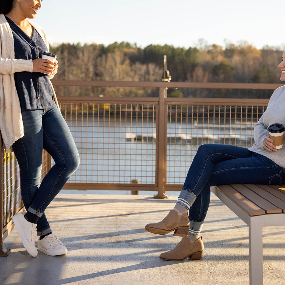 Two women on a wooden deck by a lake, one standing and one sitting on a bench, both holding coffee cups.