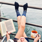 Person sitting by a body of water with legs crossed, wearing blue socks and holding a book and mug.