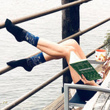 Person sitting by a waterfront holding a book, wearing blue patterned socks.