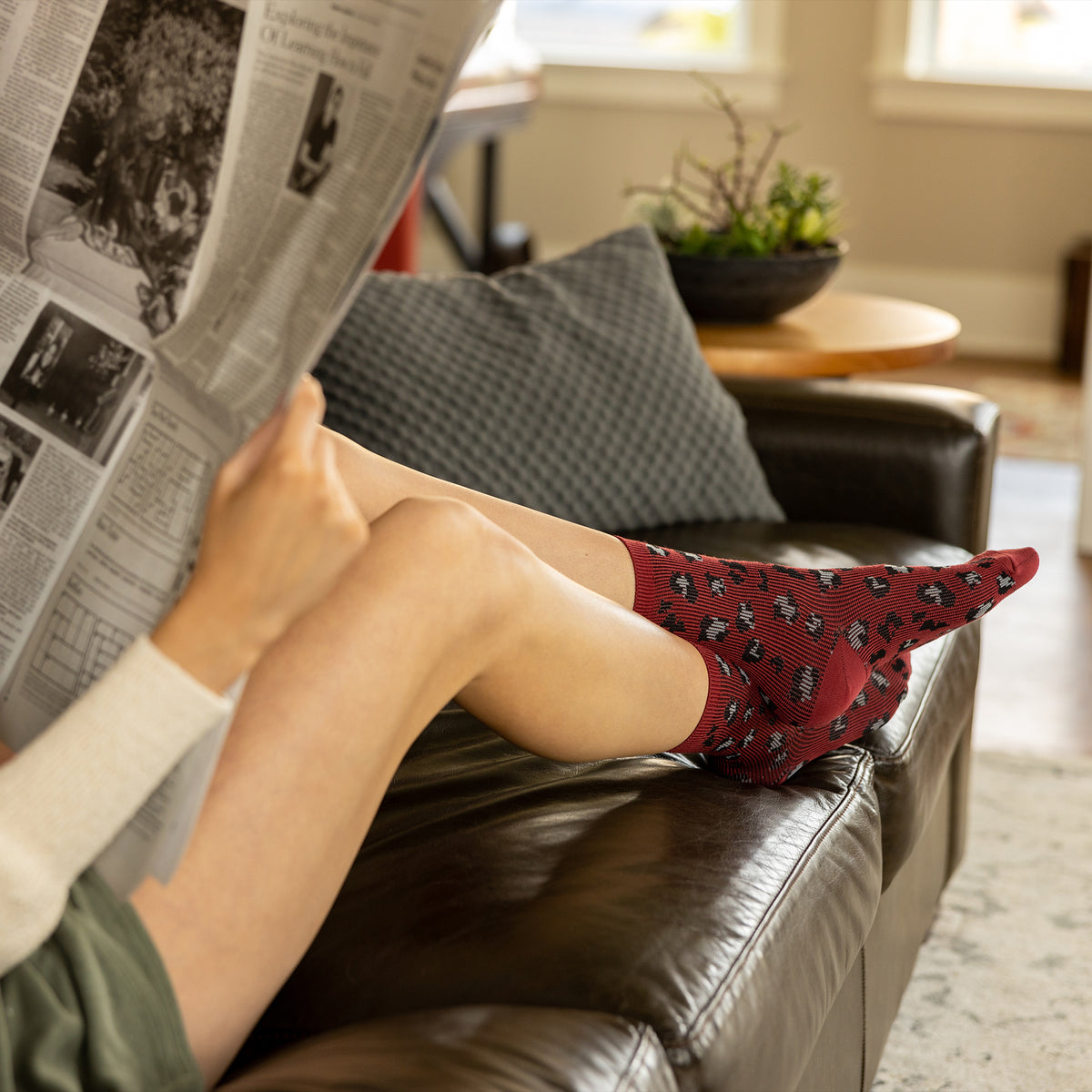 Woman sitting on a brown leather couch reading a newspaper, wearing red socks with black leopard patterns.