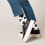 Black sneakers with white soles worn with blue jeans and cow print socks on a neutral background thumbnail