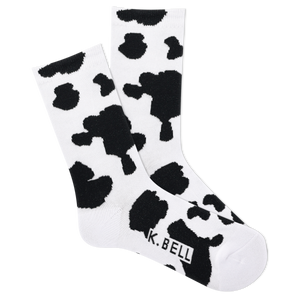 A pair of white crew socks with black cow print and the brand name 'K.BELL' written across the foot.