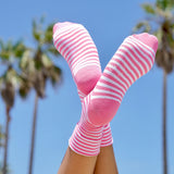 Women's pink and white striped hydrating K.Bell crew socks with aloe in front of palm trees and a clear blue sky.