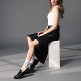 Woman sitting on a white block wearing a black skirt, white top, and black shoes with white striped socks.