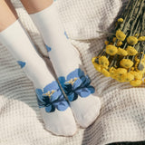 White socks with blue floral patterns held by a person next to a bouquet of yellow flowers on a textured surface.